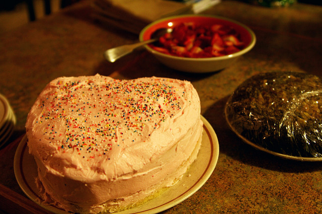 Heart-shaped Valentine cake with sprinkles. A bowl of chopped strawberries is in the background.