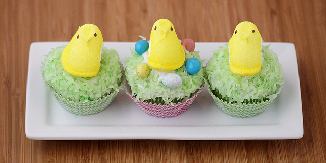 Three cupcakes on a white rectangular plate. The cupcakes are decorated with green coconut flakes and peeps. These are great alternative to traditional Easter cakes.
